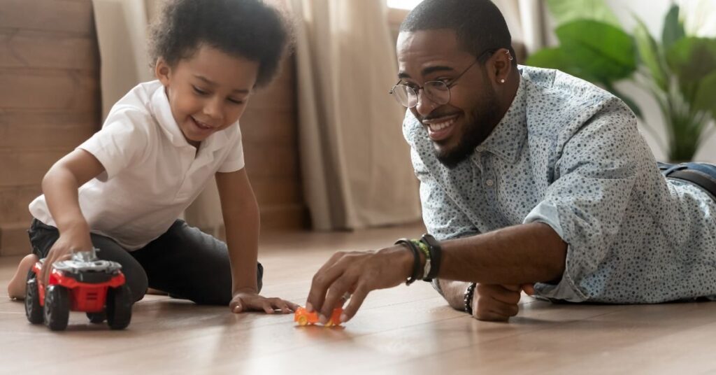 A man and a child are joyfully playing together with colorful toy cars on a carpeted floor.