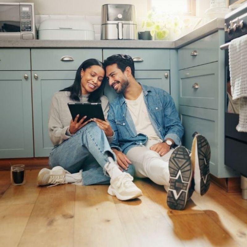 A couple sits on the kitchen floor, engaged with a tablet, surrounded by a cozy home environment.
