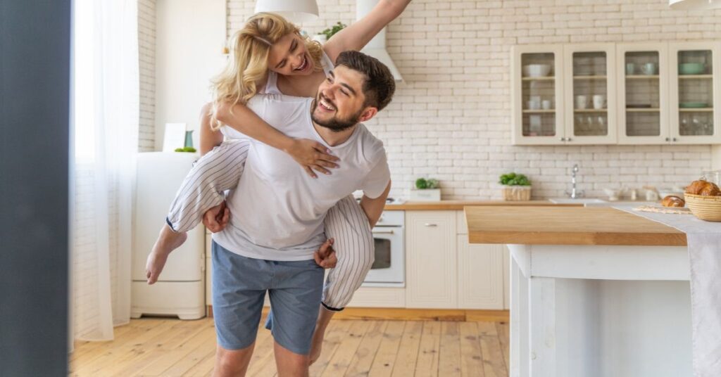 A man carries a woman in his arms, both smiling, in a cozy kitchen filled with warm lighting and homey decor.
