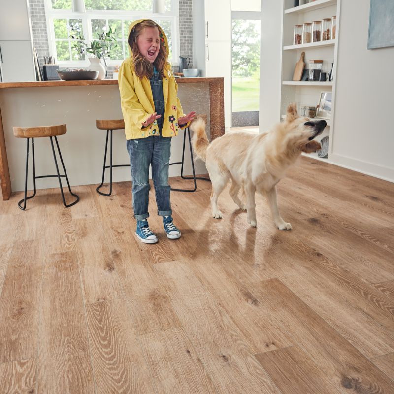 A young girl stands in a kitchen, smiling at a dog sitting beside her on the floor.

