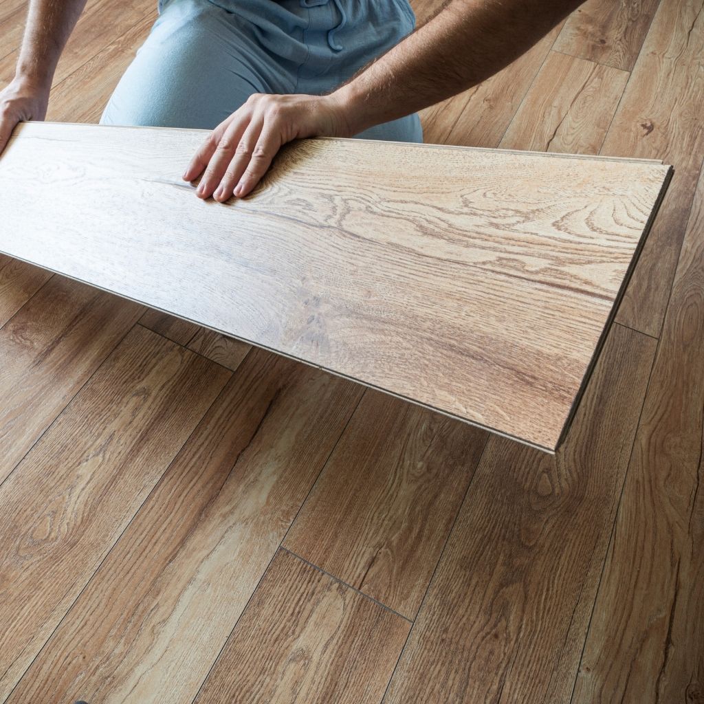 A man is positioned on the floor, holding a flooring plank